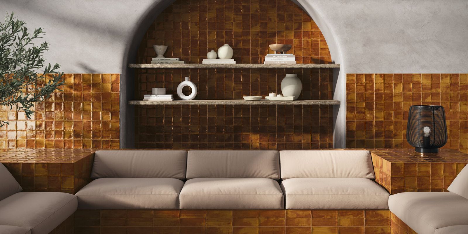 Warm-toned living space featuring glossy amber square tiles on the walls and built-in seating, with neutral beige cushions, minimalist decor on open shelves, and a sculptural arch framing the shelving.