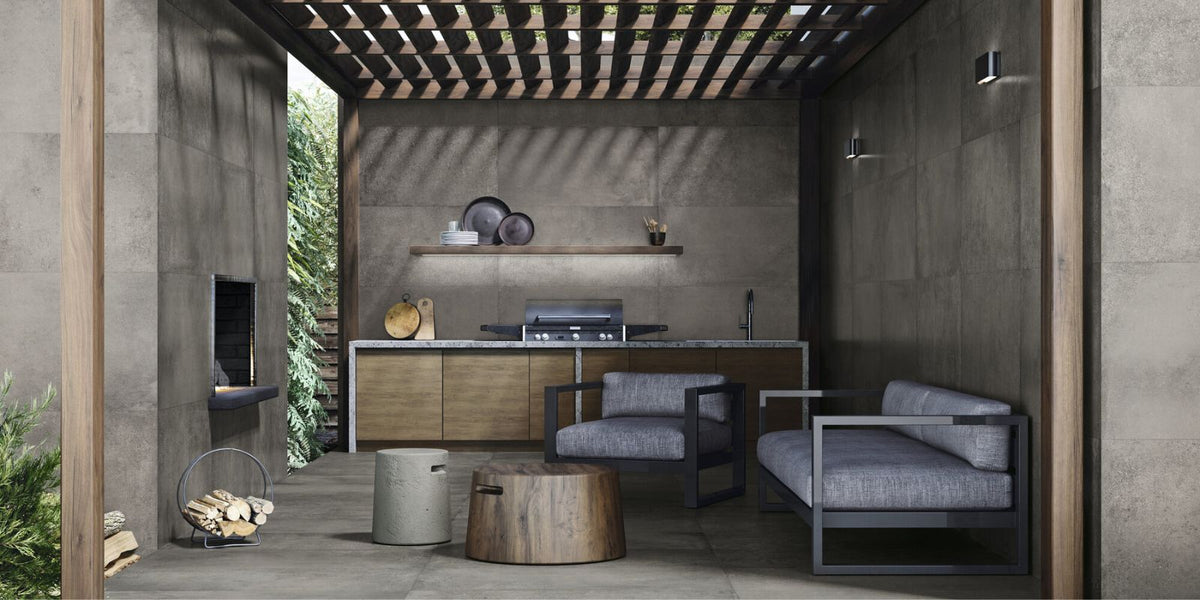 Modern outdoor kitchen and lounge area with large-format grey concrete tiles on the floor and walls, wood cabinetry, a sleek built-in grill, minimalist floating shelf, and lounge furniture with black metal frames and grey cushions, all under a wooden pergola.