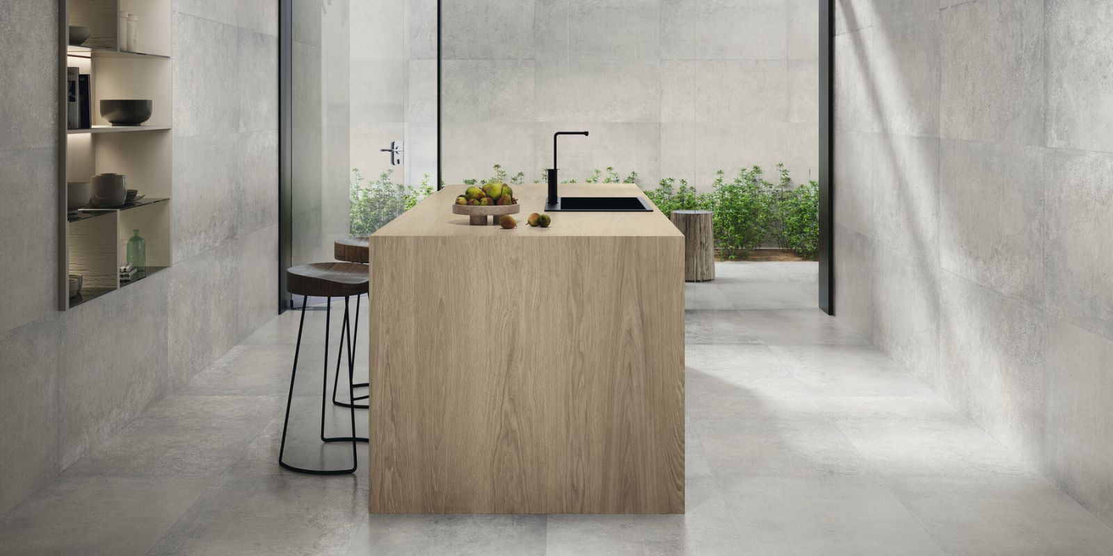 Minimalist kitchen with concrete walls and floors, a central light wood island with black fixtures, and open shelving with neutral dishware.