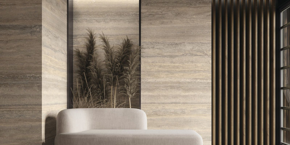 This image shows a minimalist, modern interior featuring beige-toned stone walls with subtle horizontal striations, a neutral-toned cushioned bench or seat, and decorative dried grasses displayed in a recessed wall niche. Vertical wooden slats near a window add warmth and texture, contributing to a calm, natural aesthetic.