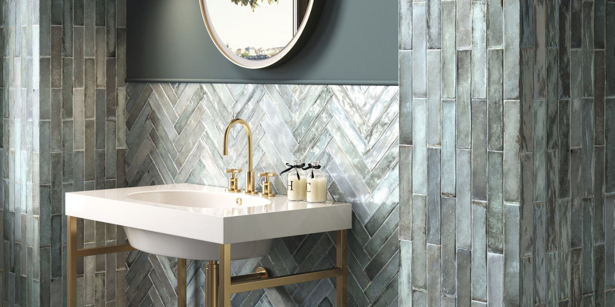 Earthy bathroom vanity area with grey and blue mosaic inspired rustic wall tiles and a free standing white sink with gold legs. 