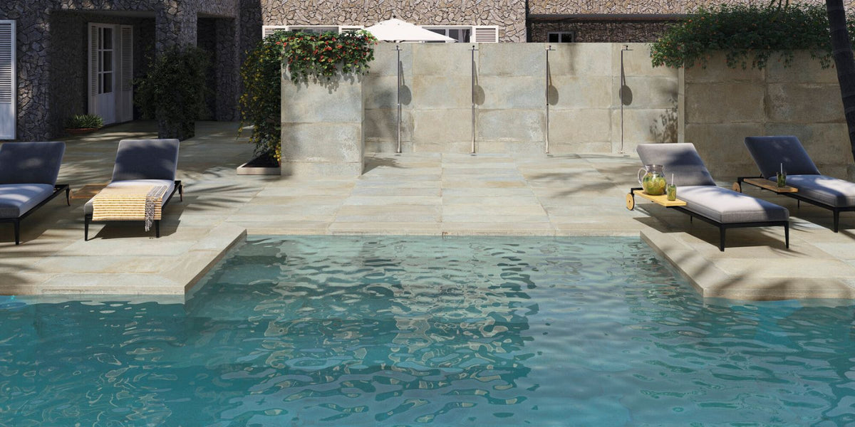 Outdoor pool with turquoise water, stone tile deck, modern gray loungers, wall showers, and stone-clad house backdrop.